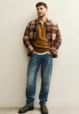 Geruite bruine en blauwe shirtjacket over een bruine trui, gecombineerd met versleten blauwe jeans en donkere bruine laarzen. Casual gelaagd outfit.