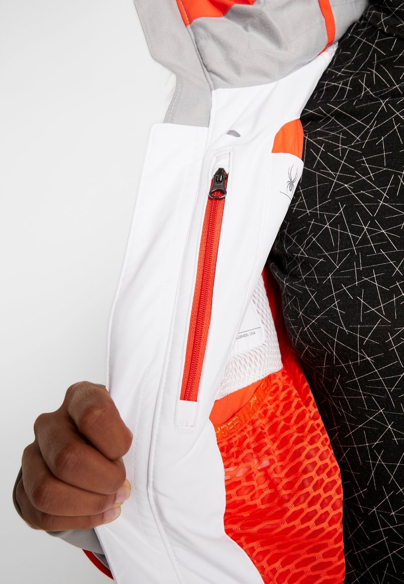 Main tenant une veste blanche et orange ouverte révélant une poche intérieure à fermeture éclair rouge et une doublure en mesh orange, portée sur une chemise noire à motifs.