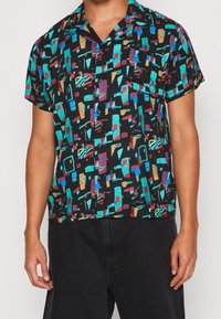 Korte mouwen shirt van zwarte stof met multicolore abstracte geometrische patronen. Voorzien van een kraag en knopen aan de voorkant.