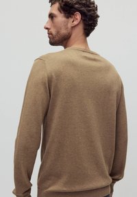 Bruine gebreide trui met een ronde halslijn. Heeft lange mouwen en ribbelmanchetten en -zoom. Zachte textuur, minimalistisch design, gedragen vanuit een zijaanzicht.
