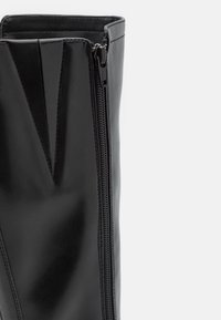 Botte noire en cuir montante jusqu'au genou avec une fermeture éclair latérale, présentant une finition lisse et des coutures verticales pour ajouter de la texture.