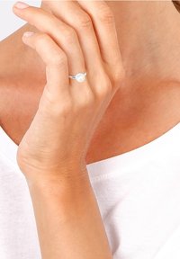 Main avec une bague en argent ornée d'une perle blanche au majeur, tenue près du visage, portant un haut blanc.