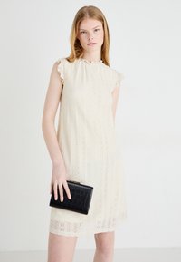 Čipkasta haljina u off-white boji s nabranim dekolteom i kratkim rukavima. Model drži teksturiranu crnu torbicu.