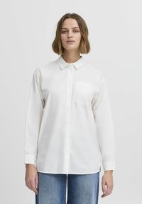 Chemise blanche à manches longues avec un col, poche poitrine simple et texture en coton doux, associée à un jean en denim.