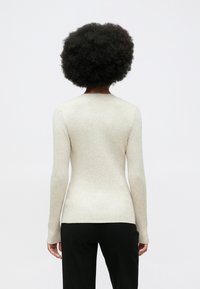 Pessoa com cabelo escuro e encaracolado, vestindo um suéter ajustado de manga longa, em bege canelado, e calças pretas, voltada de costas contra um fundo liso.