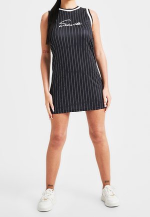Schwarzes ärmelloses Kleid mit weißen Nadelstreifen, Rundhals und Logo auf der Brust. Kombiniert mit weißen Sneakers, die ein sportliches Design aufweisen.