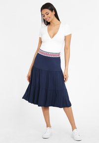 Marine tiered rok met geborduurd taille banddetail, gedragen met een korte mouw witte top en witte sneakers. Gladde stofstructuur.