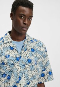 Lyhythihainen paita, jossa on vaaleansininen ja valkoinen kukkakuosi sekä sinisiä yksityiskohtia. Valmistettu kevyestä kankaasta, rento istuvuus.