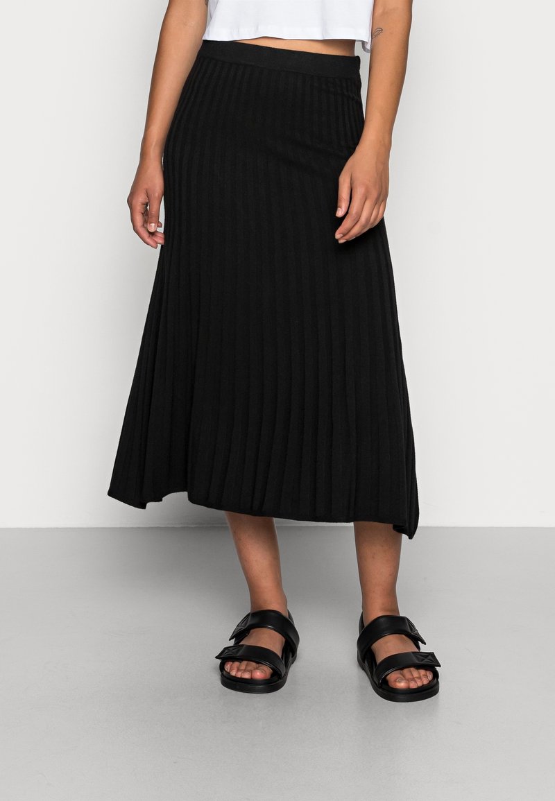 Femme portant une jupe midi plissée noire, un haut court blanc et des sandales noires à double bride, debout sur un sol gris.