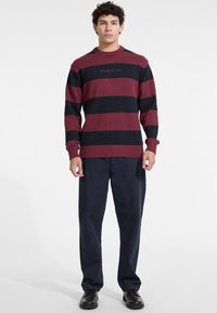 Gestreepte trui met bordeauxrode en marineblauwe horizontale strepen, ronde hals, geribbelde manchetten en onderrand, met het geborduurde "GUESS JEANS" logo aan de voorkant.