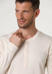 Maglietta henley a maniche lunghe color crema con texture a coste, caratterizzata da uno scollo arrotondato e tre bottoni sulla patta.