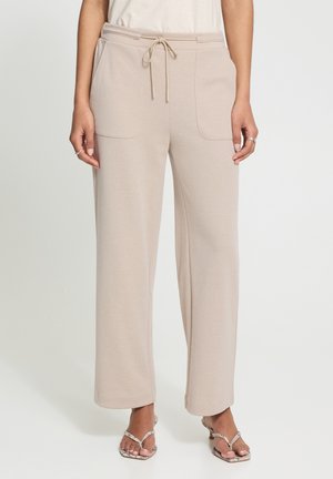 Woman wearing beige wide-leg pants with front patch pockets and drawstring waist, paired with white sandals with thin straps.