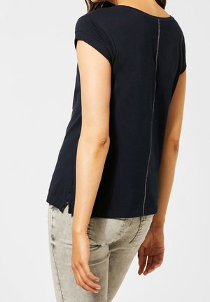 Vrouw draagt een marineblauw T-shirt met korte mouwen met zichtbare rugnaad en lichtgrijze jeans, staand met gedeeltelijk gedraaide rug.