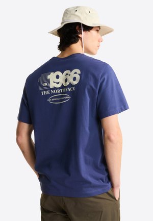 Uomo che indossa un cappello beige a tesa larga, una maglietta blu con la scritta "1966 The North Face Est. Berkeley California" e pantaloni verde oliva, con le mani nelle tasche.