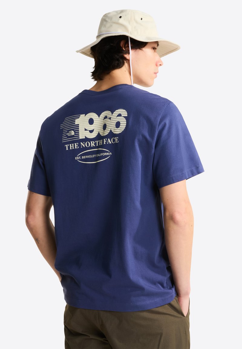 Homme portant un chapeau seau beige, un T-shirt bleu avec le texte "1966 The North Face Est. Berkeley California" et un pantalon vert olive, les mains dans les poches.