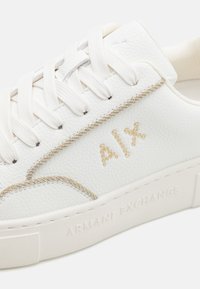 Sneakers bianchi in pelle con finitura testurizzata, dotati di dettagli dorati, lacci eleganti e "ARMANI EXCHANGE" impresso sulla suola.