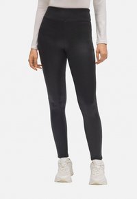 Mujer con leggings negros de cintura alta y zapatillas blancas, de pie con los brazos relajados a los lados sobre un fondo blanco.