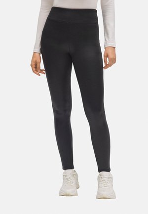 Mujer con leggings negros de cintura alta y zapatillas blancas, de pie con los brazos relajados a los lados sobre un fondo blanco.