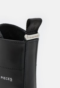 Bota de tobillo de cuero negro con textura suave, con una lengüeta de tela para tirar y un detalle metálico con la inscripción "FILLING PIECES" en el lateral.