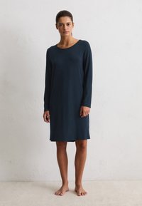 Donna in piedi a piedi nudi contro un muro bianco, che indossa un semplice abito blu scuro a maniche lunghe e lungo fino al ginocchio.