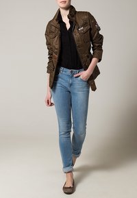 Brun vadderad jacka med flera fickor, bärs över en svart blus. Ljust blå skinny jeans och bruna ballerinas fulländar outfiten.