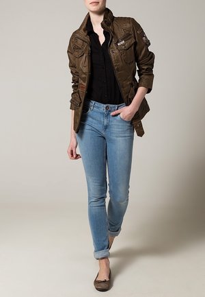 Brun vadderad jacka med flera fickor, bärs över en svart blus. Ljust blå skinny jeans och bruna ballerinas fulländar outfiten.