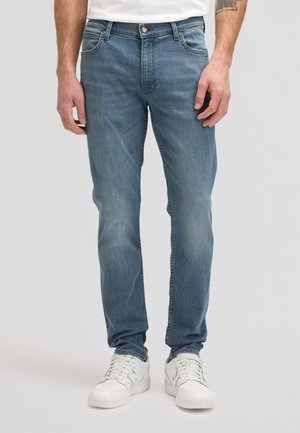 Hombre con vaqueros de pierna recta de color azul claro y zapatillas blancas, de pie contra un fondo claro y sencillo.