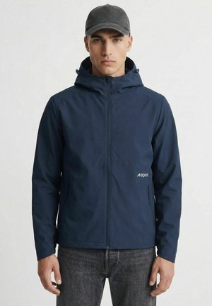 Mand iført en marineblå hættetrøje med lynlås og sort kasket, stående mod en ensfarvet lys baggrund.