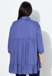 Blusa di cotone viola con design arricciato, maniche a tre quarti e orlo a volant. Presenta un colletto e una texture morbida.