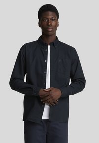 Camisa de botones azul marino de algodón, con collar tipo button-down, bolsillo en el lado izquierdo del pecho y camiseta blanca debajo. El modelo está de pie con los brazos cruzados.
