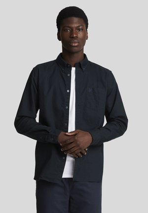 Camisa de botones azul marino de algodón, con collar tipo button-down, bolsillo en el lado izquierdo del pecho y camiseta blanca debajo. El modelo está de pie con los brazos cruzados.