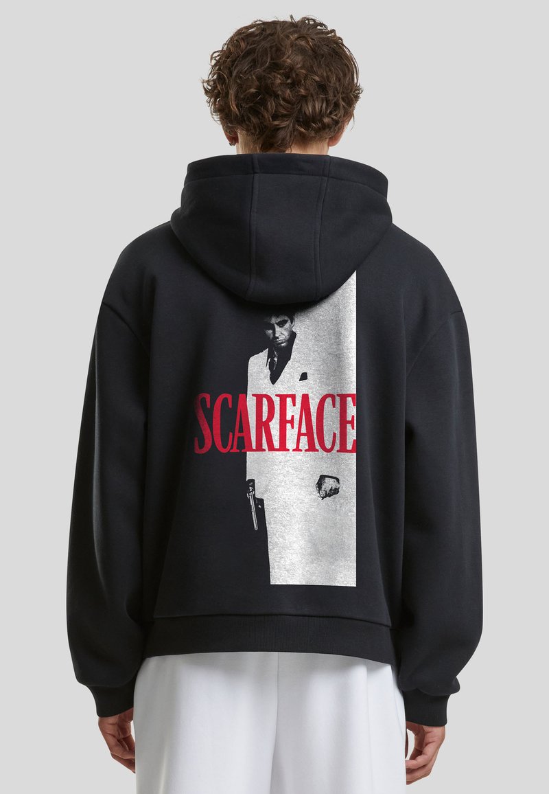 Personne aux cheveux bouclés portant un sweat à capuche noir montrant au dos une affiche du film Scarface en noir, blanc et rouge.