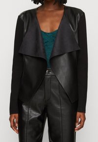 Schwarze Kunstlederjacke mit vorderer Drapierung, mattem Finish und langen Ärmeln. Getragen über einem türkisfarbenen Spitzenoberteil.