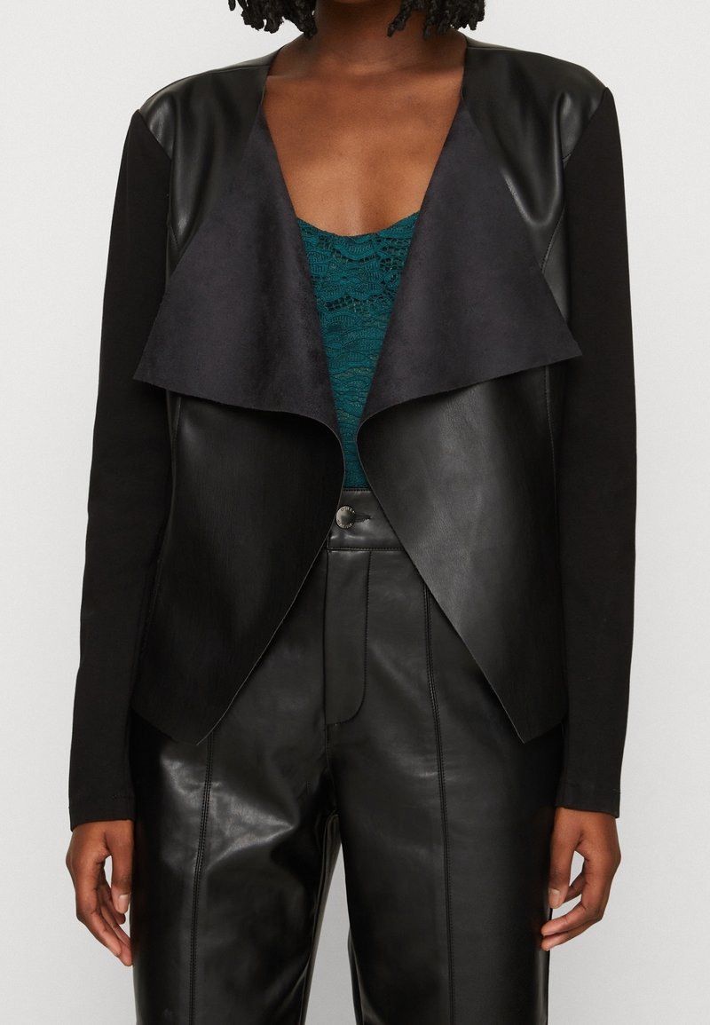 Schwarze Kunstlederjacke mit vorderer Drapierung, mattem Finish und langen Ärmeln. Getragen über einem türkisfarbenen Spitzenoberteil.