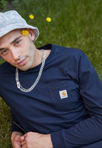 Tengerészkék hosszúujjú ing mellzsebbel és Carhartt logóval, ezüstlánc nyaklánccal és világosszürke bőrsapkával, zöld fű előtt.