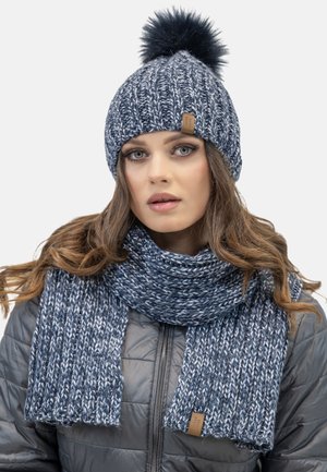 Blauer gestrickter Beanie mit Pompon und passendem Schal, ausgestattet mit einer groben Rippstruktur und einem Lederschild.