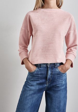 Frau trägt ein rosa geripptes Langarmshirt, das in hoch taillierte blaue Jeans gesteckt ist, die Hände in den Taschen, vor einem einfarbigen Hintergrund.