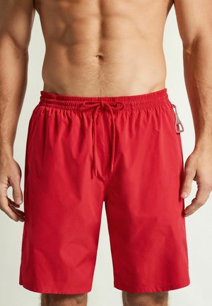 Homme portant un short de bain rouge avec taille élastique et cordon, torse visible de la moitié de la poitrine jusqu'aux genoux sur fond uni.