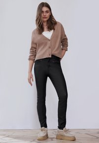 Cárdigan marrón claro con tres botones, usado sobre una camiseta blanca, combinado con jeans skinny negros y zapatos de piel de pelo beige.