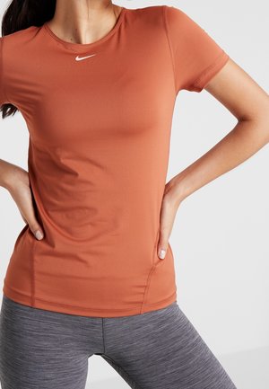 Kobieta ubrana w pomarańczową sportową koszulkę Nike z krótkim rękawem i szare legginsy treningowe, z rękami opartymi na biodrach, na jednolitym tle.