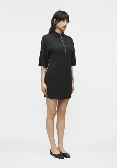 Γυναίκα που στέκεται φορώντας μαύρο φόρεμα μήκους μέχρι το γόνατο με μισά μανίκια, γιακά, φερμουάρ μπροστά και μαύρα μπαλαρίνα παπούτσια, μπροστά σε απλό ανοιχτό φόντο.