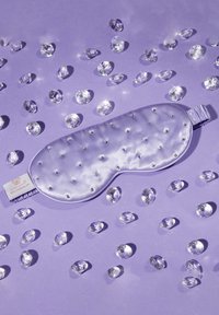 Maschera da sogno in raso viola chiaro con borchie argento, bordi arrotondati e una fascia elastica, circondata da pietre simili a gemme trasparenti sparse.