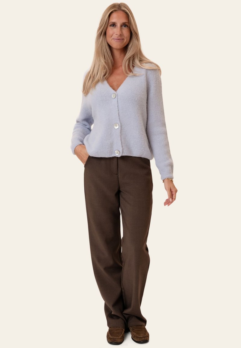Femme aux longs cheveux blonds portant un cardigan bleu clair boutonné, un pantalon marron et des chaussures marron, debout avec une main dans la poche.
