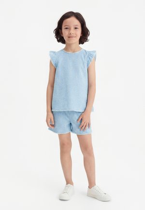 Child standing, wearing blue and white patterned sleeveless top and matching shorts with white sneakers, smiling at camera.