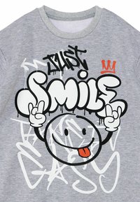 Grijs t-shirt met zwart-witte graffiti-stijl tekst, een lachend cartoon gezicht, rood kroontje accent en speelse handgebaren.