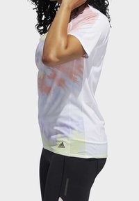 Femme aux cheveux bouclés portant un t-shirt tie-dye blanc Adidas et des leggings noirs, debout de profil avec la main près du visage.