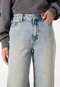 Jeans taille haute bleu clair avec un effet délavé, présentant des poches avant et une fermeture à bouton. Le matériau semble être du denim avec une texture douce.