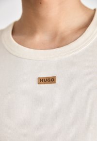 Off-white rebrované tričko z látky s hnedou koženou nášivkou s embosovaným nápisom "HUGO". Úzky výstrih s jemným textúrnym detailom.
