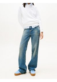 Sudadera blanca de cuello redondo, camisa de botones azul clara debajo, jeans de pierna ancha azul clara y zapatillas azul claras con suelas marrones.