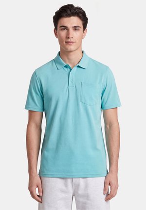 Junger Mann trägt ein hellblaues kurzärmeliges Polohemd mit Brusttasche und helle Shorts vor einem einfarbigen Hintergrund.